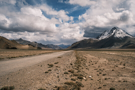 View of the Pamir Mountains rising above a vast arid plain with a dirt road leading towards the snow-capped peaks under a dramatic cloudy sky in Jirgatol, Gorno-Badakhshan Auto, Tajikistan.