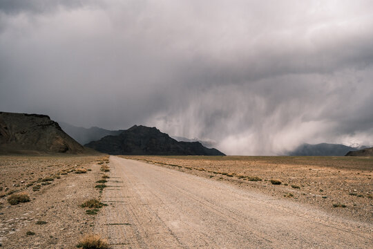 View of the rugged Pamir Mountains landscape with a desolate dirt road stretching into the distance under a dramatic stormy sky with rain showers in Jirgatol, Gorno-Badakhshan Auto, Tajikistan.