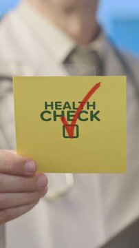 Medical professional in a white lab coat with a stethoscope holding up a yellow sticky note that says health check with a large red check mark, symbolizing preventive care and medical examination