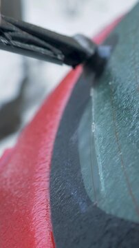 Male hand lifts rear wiper on red vehicle and removes brittle ice crust from blade. Frozen residue sticks to rubber, weakens contact with glass. Severe cold, icy street create hazardous conditions