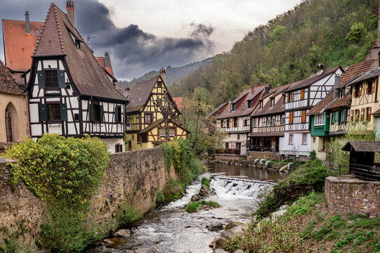 Dark cloud tears open above the wooded ridge behind Kaysersberg while the Weiss river runs over a weir between a tall black and white gable and a terrace of riverside houses.
