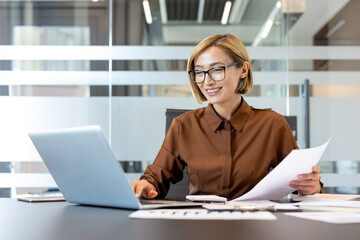 Professional businesswoman smiling and looking at a laptop screen while holding paperwork and...