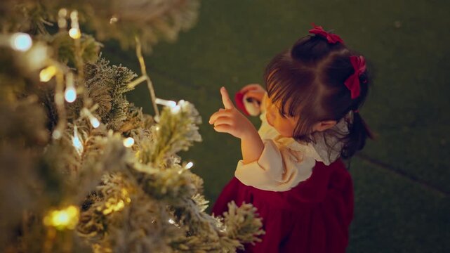 Cute Asian toddler girl in red dress pointing at glowing Christmas tree lights with curiosity, little child enjoying festive holiday decoration, Christmas celebration, childhood wonder