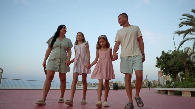 Four family members enjoy a walk together along the beach during sunset. They hold hands and share smiles as they stroll on a path lined with palm trees.