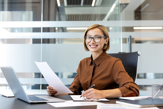 Professional businesswoman sitting at a desk in a contemporary office, smiling confidently while holding a document and writing on a clipboard, surrounded by paperwork and a laptop