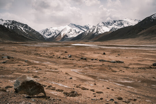 View of the Pamir Mountains with snow-capped peaks rising above an arid, rocky landscape under a cloudy sky in Jirgatol, Gorno-Badakhshan Auto, Tajikistan.