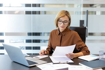 Fototapeta na wymiar Professional woman at modern office desk reviewing financial documents and using a calculator and laptop to manage budgeting, taxes and accounting with focused concentration