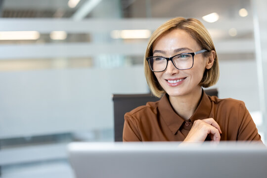 Smiling businesswoman in eyeglasses and a brown blouse works confidently at a laptop in a bright modern office, looking to the side with a focused, optimistic expression