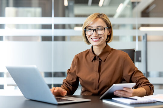 Young professional businesswoman smiling at camera, sitting at office desk, successfully managing tasks and responsibilities while using a laptop and a digital tablet for efficient workflow