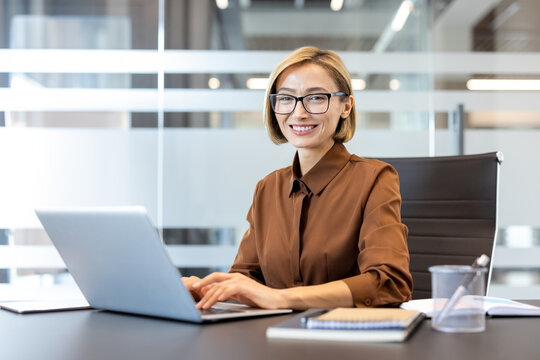 Smiling businesswoman sitting at her desk, wearing eyeglasses, using a laptop in a contemporary office environment, confidently managing tasks and representing professional success