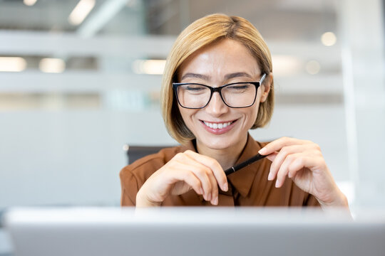 Cheerful businesswoman wearing glasses and a brown shirt, looking down and smiling while holding a pen, focused on her work at a laptop in a bright corporate office setting