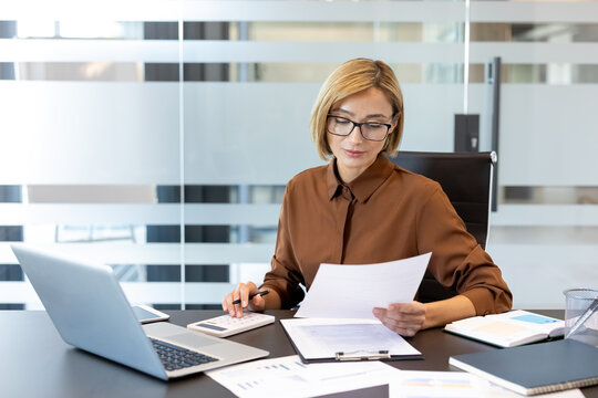 Professional woman at modern office desk reviewing financial documents and using a calculator and laptop to manage budgeting, taxes and accounting with focused concentration