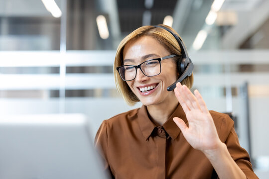 Smiling woman wearing a headset with a microphone and glasses, communicating virtually on a laptop from a modern office, symbolizing remote work, online support, and digital communication