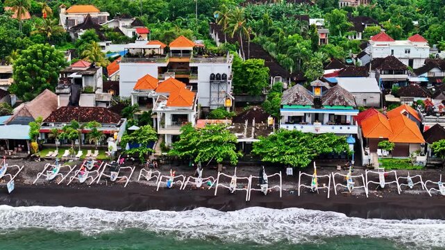 Aerial view of Amed Beach featuring traditional outrigger boats on the black sand shore with lush tropical vegetation and coastal buildings in Bali, Indonesia.