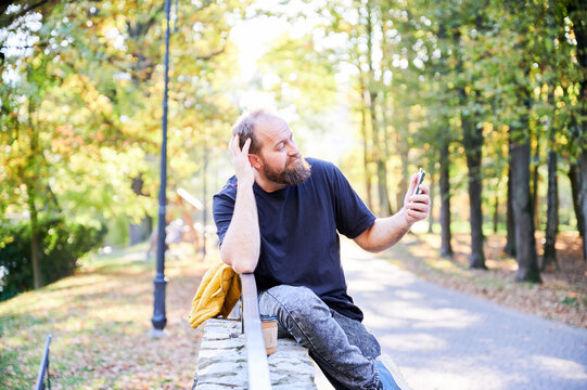 Man selfies casually. Relaxed man with phone picture. Casual portrait of man using phone outdoors during leisure. Individual posing with phone while leaning on park bench in casual manner