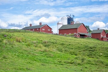 Red barns and silos on a grassy hill under a blue sky © Maxwell
