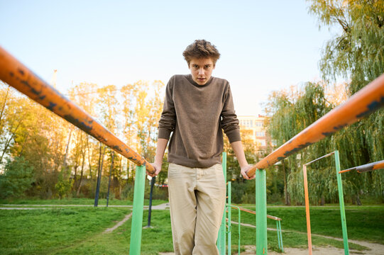 Teen caucasian male gripping parallel bars outdoors, focused stance with shoulders engaged and steady gaze. Autumn light highlights textured sweater and urban park equipment, grassy paths