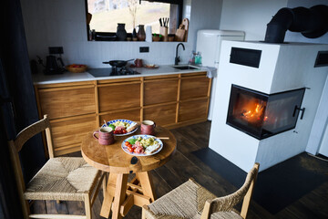 Obraz premium Cozy kitchen interior with fireplace and wooden dining table set for breakfast, plates of healthy food and coffee, concept of hygge lifestyle, home comfort, warm atmosphere