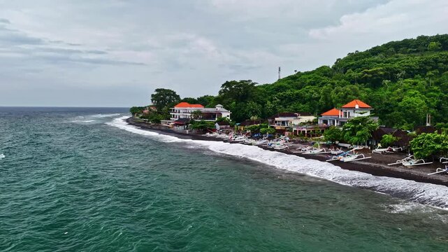 Aerial view of Amed Beach featuring black sand, traditional outrigger boats, and lush green hills along the coastline in Bali, Indonesia.
