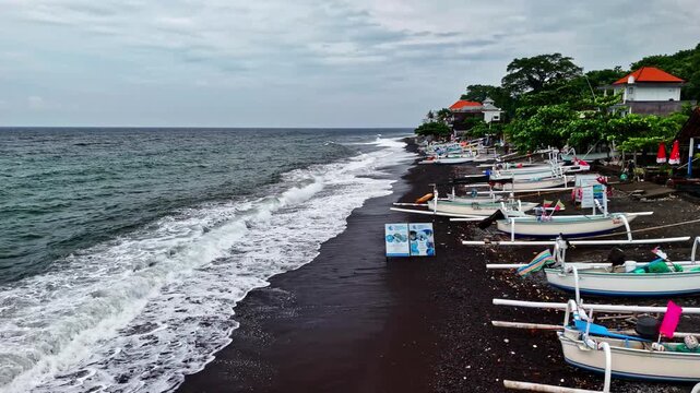 Aerial view of Amed Beach featuring traditional outrigger boats resting on the black sand shore with ocean waves and coastal buildings in Bali, Indonesia.