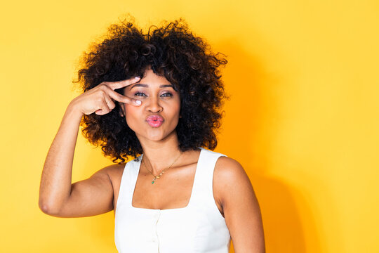 young latin woman making a peace sign gesture over her eye, puckering her lips playfully against a bright yellow background, expressing fun and confidence