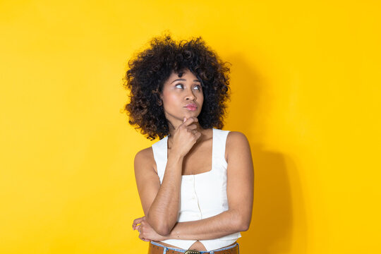 Thoughtful African American woman with a pensive expression, hand on chin, looking sideways against a bright yellow background with copy space