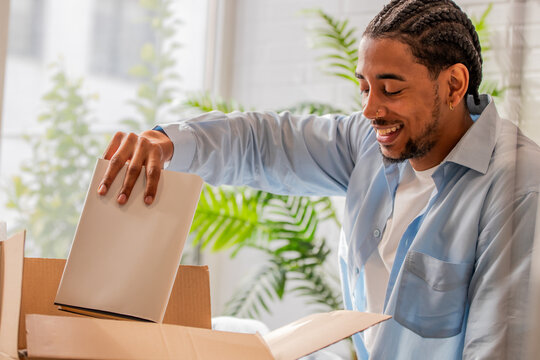 smiling young black man unpacking a book from a cardboard box while moving into his new home, surrounded by houseplants and natural light