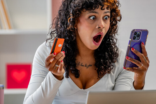 shocked mixed race woman with wide eyes and open mouth holding a credit card and smartphone, expressing surprise and disbelief while looking at her phone screen
