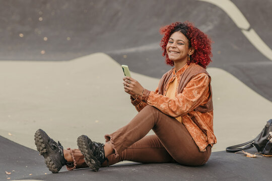 Young hispanic black woman sitting outdoors at a skate park, focused expression, using a smartphone
