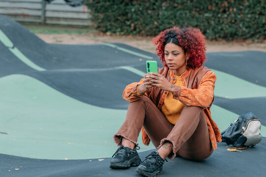Young mixed-race woman sitting outdoors at a skate park, focused expression, using a smartphone