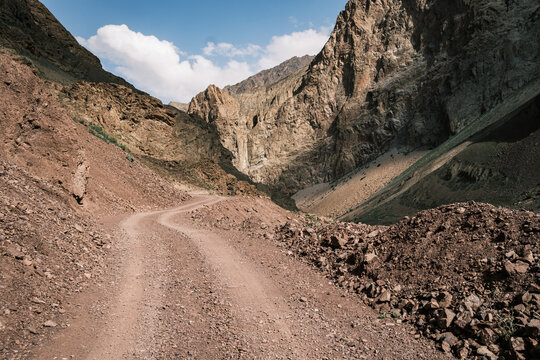 View of the rugged dirt road winding through the Bartang valley surrounded by steep rocky mountains and arid slopes in Chah Ab, Gorno-Badakhshan Auto, Tajikistan.