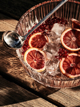 Ribbed glass punch bowl filled with sparkling pomegranate drink blood orange slices aril ice silver ladle wooden table sunlight copy space