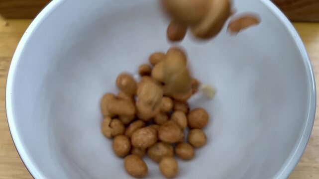 Closeup of roasted peanuts falling into a white bowl on a wooden table. The shot captures the movement and texture of the nuts as they collect inside the bowl.