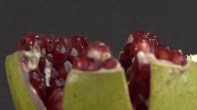 Closeup of cut pomegranate showing glossy red aril inside pale green peel. Macro view highlights the juicy fruit texture and natural color contrast against a dark background.