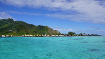 Fototapeta premium A wide, stunning view across the vibrant turquoise waters of a lagoon in Moorea, French Polynesia, showing overwater resort bungalows at the base of dramatic green volcanic hills