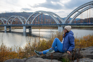 Woman wearing blue casual sportswear and a beige knit beanie rests on rocks above Jezioro Mucharskie with the multi-arch bridge behind her. The composition combines autumn travel, waterside recreation © Volha