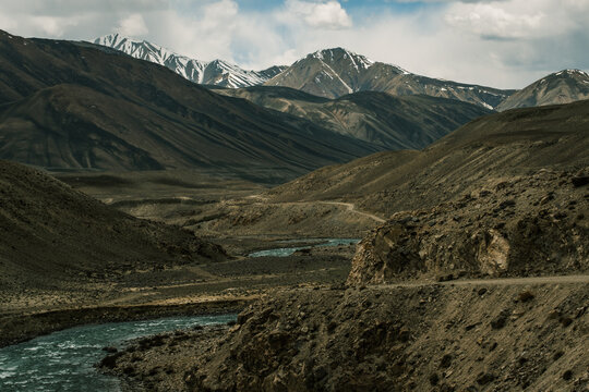 View of the Wakhan Corridor featuring a winding river flowing through a rugged mountain valley with snow-capped peaks under a cloudy sky in Badakhshan, Afghanistan.