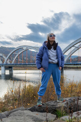 Full-length portrait of an adult woman standing confidently on shoreline rocks overlooking Jezioro Mucharskie and its monumental arched bridge. Blue puffer jacket, joggers, sneakers and sunglasses cre © Volha