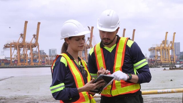 Dockworkers Collaborate: Two industrious dockworkers, clad in hard hats and high-visibility vests, confer over a tablet amidst a busy shipping port, overseeing maritime operations and logistics.