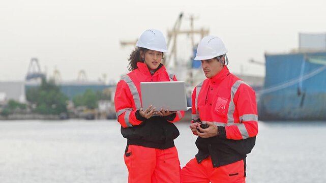 Port Professionals: Two industrious port workers, donned in high-visibility safety gear and hard hats, collaborate intently over a laptop amidst the backdrop of maritime operations and docked vessels.