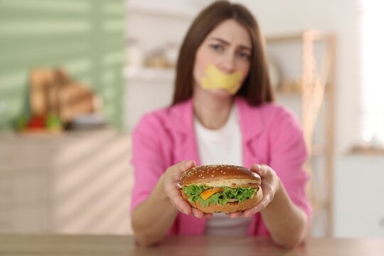 Diet. Woman with taped mouth holding hamburger indoors, selective focus. Space for text