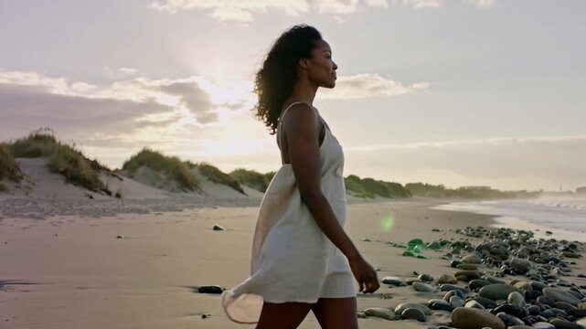 Walking woman starting midstep along rocky beach at sunrise, thinking in light sundress and bangle