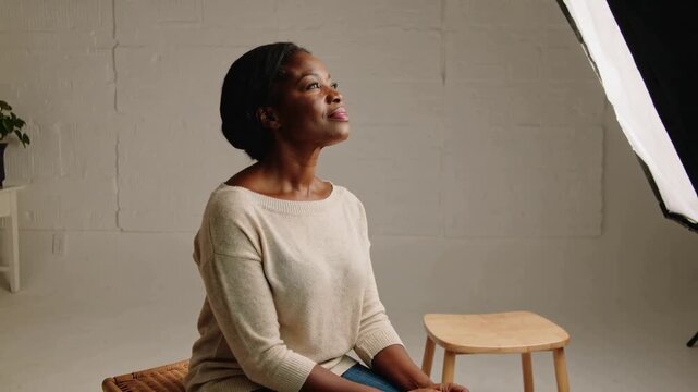 Responding to softbox shining, woman in beige sweater and blue jeans straightening on woven stool