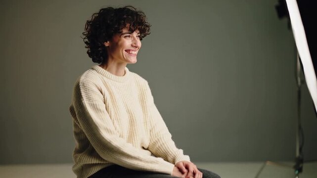 Turning male model wearing cream sweater on stool in studio, reacting to softbox light, laughing