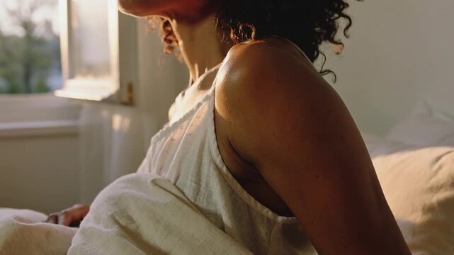 Sunlight drawing woman in camisole raising torso and gazing at open window, hand by blanket, mug
