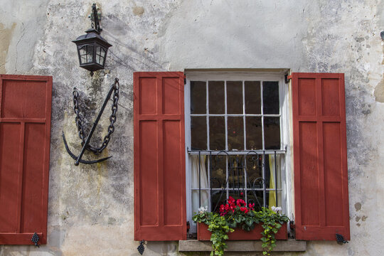 View of a historic building facade with red shutters, a flower box, a wall lantern, and a decorative anchor on the stucco wall, Charleston, South Carolina, United States.