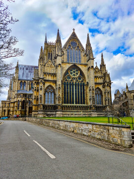 View of Lincoln Cathedral featuring its intricate Gothic architecture, stained glass windows, and stone facade under a cloudy blue sky in Lincoln, England, United Kingdom.