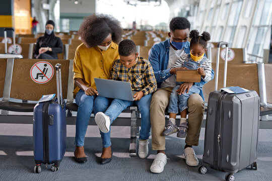 A Black family with two children sits in an airport terminal, wearing face masks and using a laptop and tablet while waiting for their flight