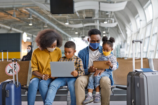 A Black family sits together in an airport terminal, all wearing protective face masks while using electronic devices and surrounded by their travel bags