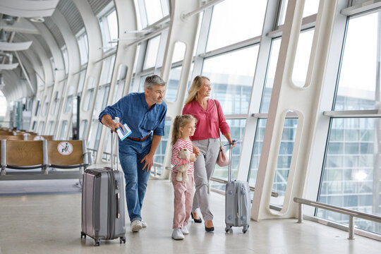 A happy family with suitcases walks through a modern airport terminal with large windows overlooking the tarmac